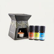 A Tanamera essential oil diffuser and burner set on a clean white background, featuring a dark gray ceramic diffuser with gold geometric patterns and a lit tealight candle, alongside three small amber bottles of essential oils.